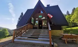 Drewniana bacówka pod honem, z kamiennymi schodami i drewnianymi ławami, otoczona zielenią.
