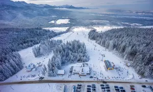 Zimowy krajobraz z trasami narciarskimi i lasem iglastym. W oddali góry. Widoczna bacówka Małe Ciche z narciarzami i parkingiem.