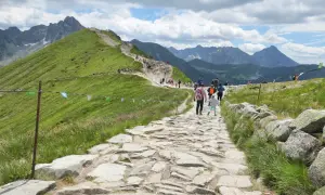Kamienista ścieżka na zielonym zboczu górskim z turystami i widokiem na Tatry.