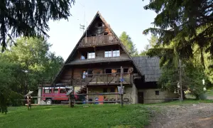 Drewniana bacówka pod Małą Rawką, z czerwonym Land Roverem zaparkowanym obok. Otoczona zielenią i drzewami.