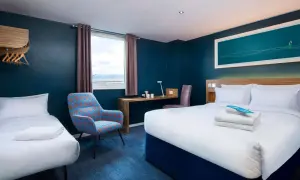 Pokój hotelowy z dwoma łóżkami, biurkiem i fotelem. Zastanawiasz się, ile kosztuje nocleg w Londynie?