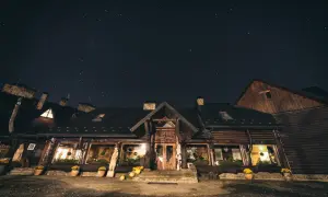 Nocne niebo pełne gwiazd nad drewnianą bacówką Biały Jeleń. Ciepłe światło sączy się z okien, zapraszając do środka.