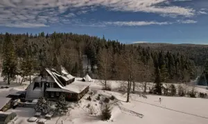 Zimowa sceneria wokół bacówki nad Wierchomlą. Śnieg pokrywa dachy, drzewa i okoliczne wzgórza.