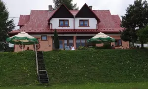 Urocza bacówka na zadzielu z czerwonym dachem, zielonymi parasolami i ludźmi odpoczywającymi na tarasie.