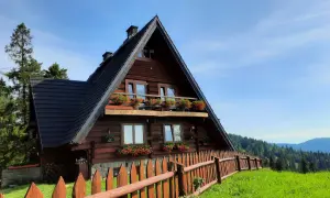 Drewniana bacówka na Maciejowej, otoczona zielenią i drewnianym płotem, z balkonem pełnym kwiatów.