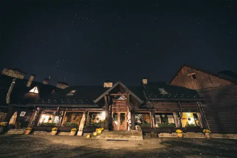 Nocne niebo pełne gwiazd nad drewnianą bacówką Biały Jeleń. Ciepłe światło sączy się z okien, zapraszając do środka.