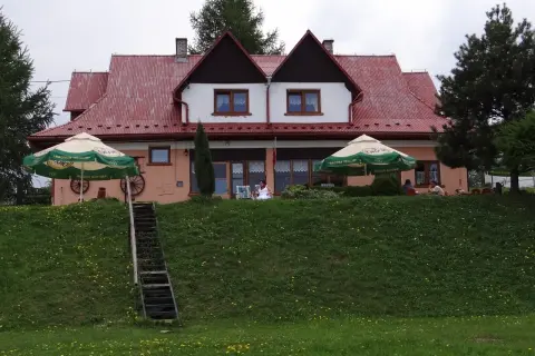 Urocza bacówka na zadzielu z czerwonym dachem, zielonymi parasolami i ludźmi odpoczywającymi na tarasie.