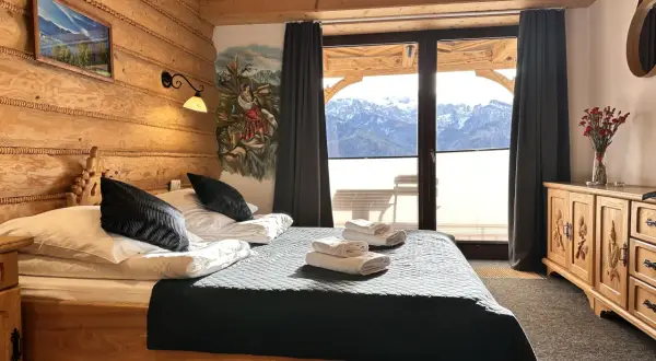 Przytulna sypialnia w stylu góralskim w Willi Kominiarski Wierch. Drewniane ściany, łóżko z czarną narzutą i widok na Tatry.