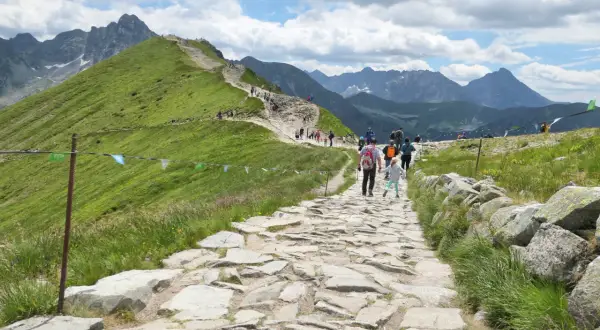 Kamienista ścieżka na zielonym zboczu górskim z turystami i widokiem na Tatry.