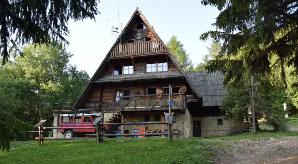 Drewniana bacówka pod Małą Rawką, z czerwonym Land Roverem zaparkowanym obok. Otoczona zielenią i drzewami.