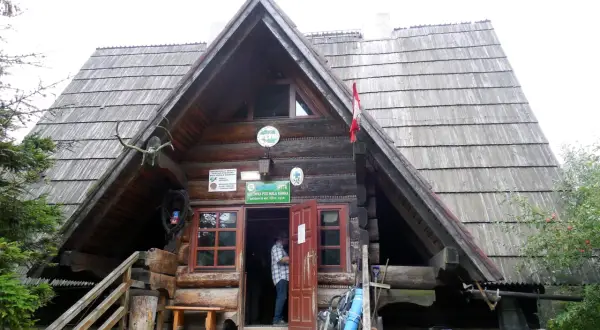 Drewniana bacówka mała rawka z charakterystycznym dachem. Przed wejściem stoi mężczyzna, a obok niego plecaki i mata.