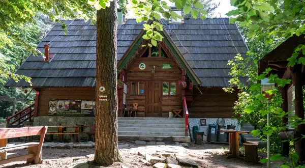 Drewniana bacówka PTTK w Bartnem, otoczona zielenią, zaprasza do odpoczynku. Na ścianie wisi tablica informacyjna.