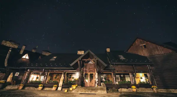 Nocne niebo pełne gwiazd nad drewnianą bacówką Biały Jeleń. Ciepłe światło sączy się z okien, zapraszając do środka.