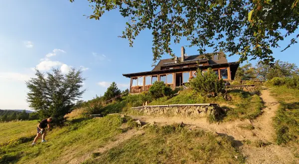 Bacówka PTTK nad Wierchomlą, drewniany dom z tarasem, otoczony zielenią, na zboczu wzgórza.