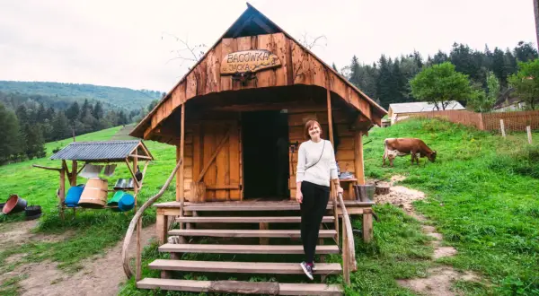 Kobieta przed bacówką u Jancoka, gdzie można kupić oscypki. W tle pasąca się krowa i górski krajobraz.