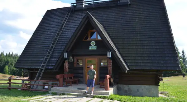Mężczyzna stoi przed drewnianą bacówką Krawców Wierch. Dach kryty gontem, pochyła drabina.