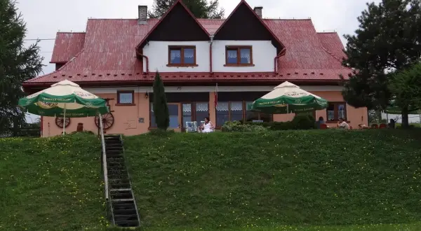 Urocza bacówka na zadzielu z czerwonym dachem, zielonymi parasolami i ludźmi odpoczywającymi na tarasie.