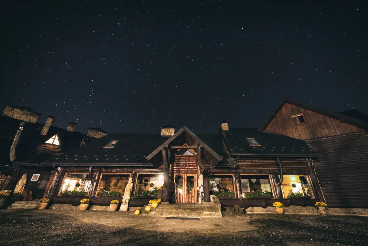 Nocne niebo pełne gwiazd nad drewnianą bacówką Biały Jeleń. Ciepłe światło sączy się z okien, zapraszając do środka.