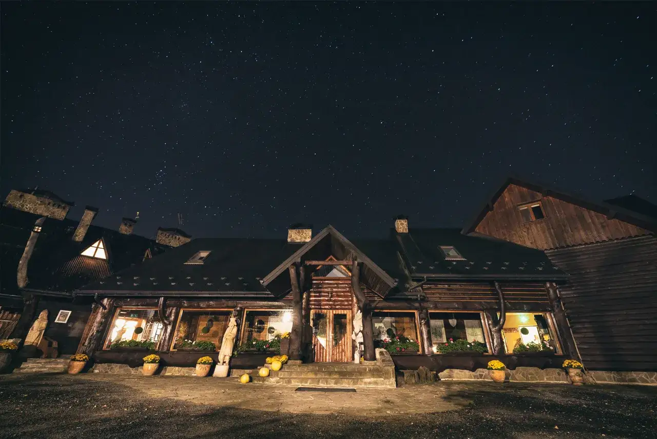 Nocne niebo pełne gwiazd nad drewnianą bac&oacute;wką Biały Jeleń. Ciepłe światło sączy się z okien, zapraszając do środka.