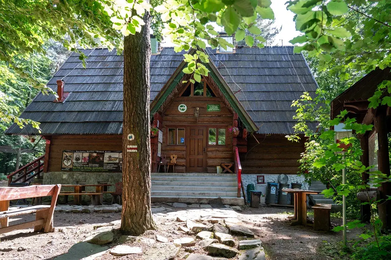 Drewniana bacówka PTTK w Bartnem otoczona zielenią. Przed schodami ławki i stoły, a obok drzewo z oznaczeniem szlaku.
