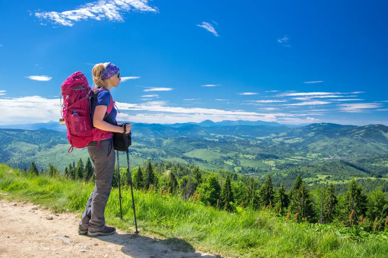 Kobieta z plecakiem i kijkami trekkingowymi podziwia widok na góry. Zastanawia się, co spakować w góry na taką wyprawę.