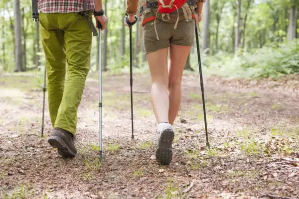 Para wędrówki w góry latem: mężczyzna w zielonych spodniach, kobieta w krótkich spodenkach, oboje z kijkami trekkingowymi, idą leśną ścieżką.