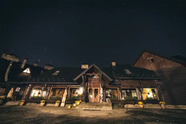 Nocne niebo pełne gwiazd nad drewnianą bacówką Biały Jeleń. Ciepłe światło sączy się z okien, zapraszając do środka.
