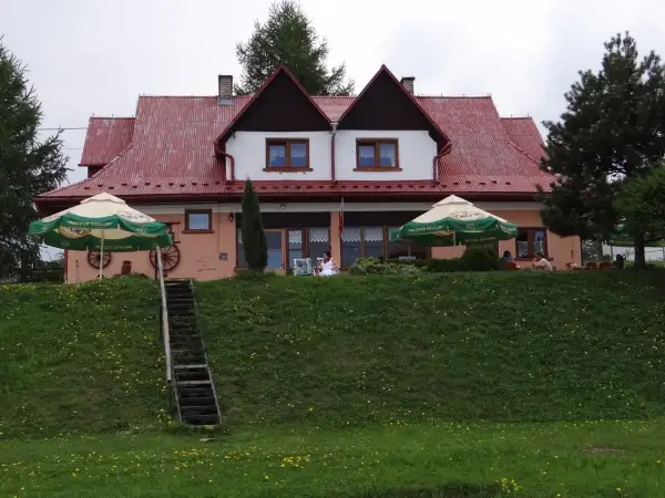 Urocza bacówka na zadzielu z czerwonym dachem, zielonymi parasolami i ludźmi odpoczywającymi na tarasie.